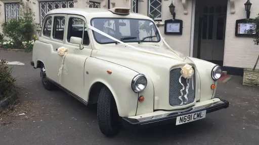 Old English White classic taxi cab decorated with white wedding ribbons, parked outside a venue with buildings in the background
