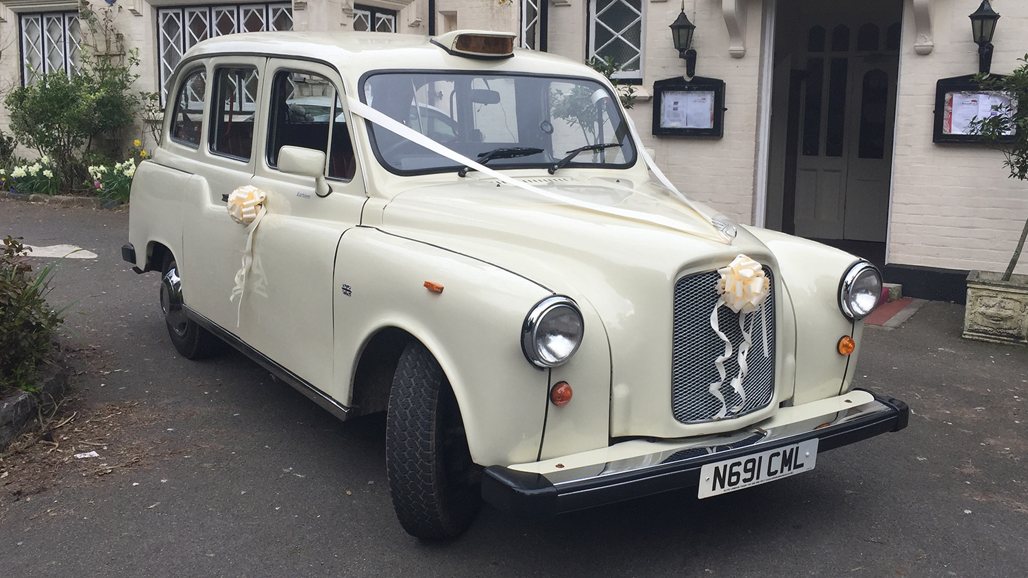 Old English White classic taxi cab decorated with white wedding ribbons, parked outside a venue with buildings in the background