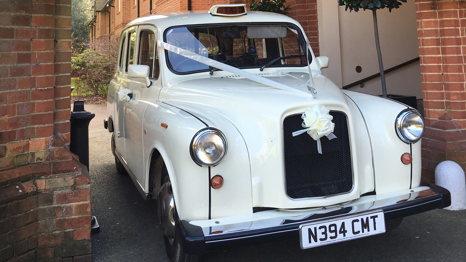 Front view of a classic taxi cab in Old English White with wedding ribbons on the grille, parked near a brick building.