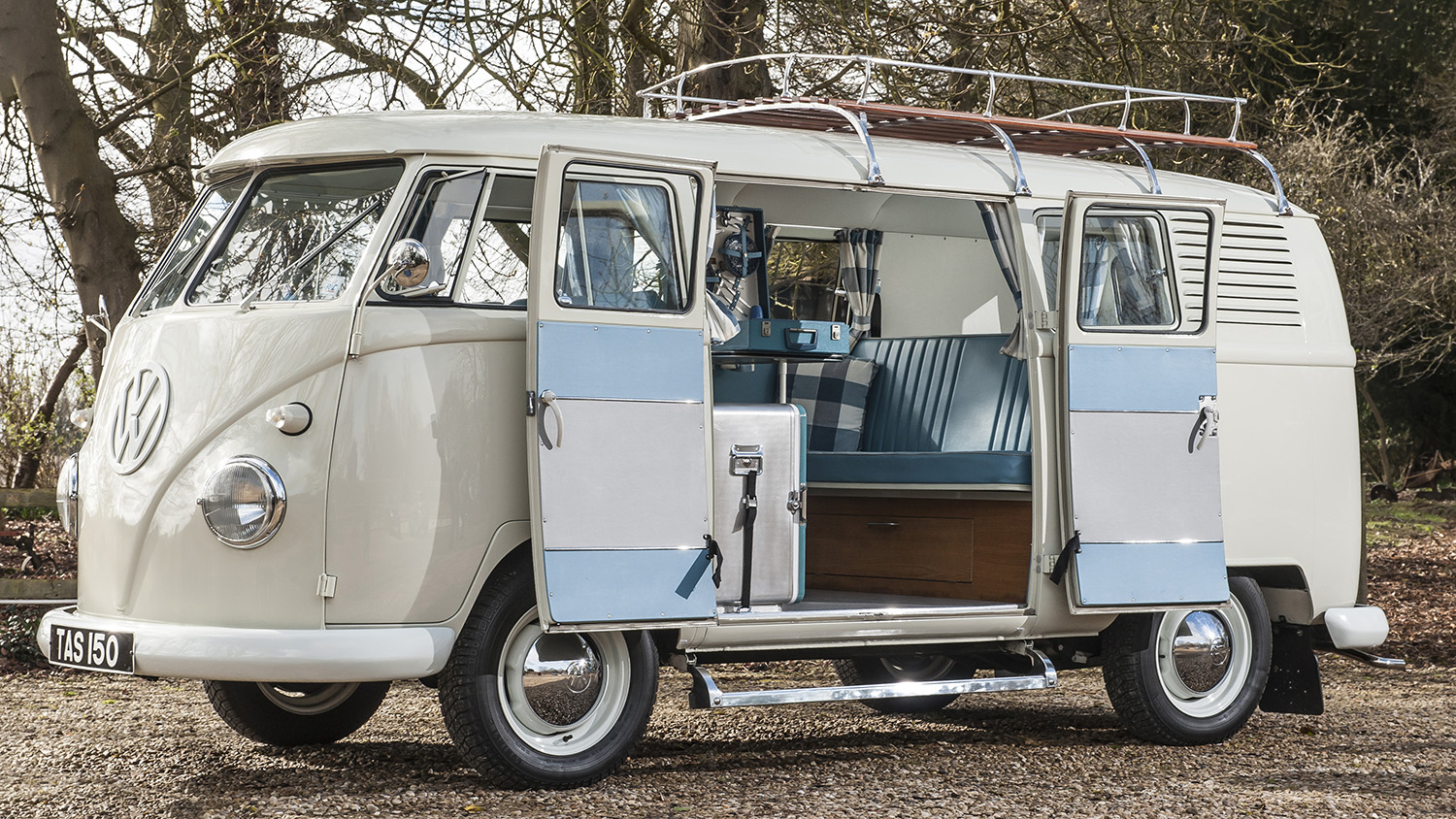 Cream VW split screen campervan with roof rack parked in a woodland setting, side door open revealing turquoise interior.