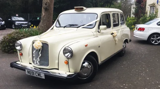 Front three-quarter view of an Old English White classic taxi cab parked on a driveway, chrome grille and rounded classic styling visible.