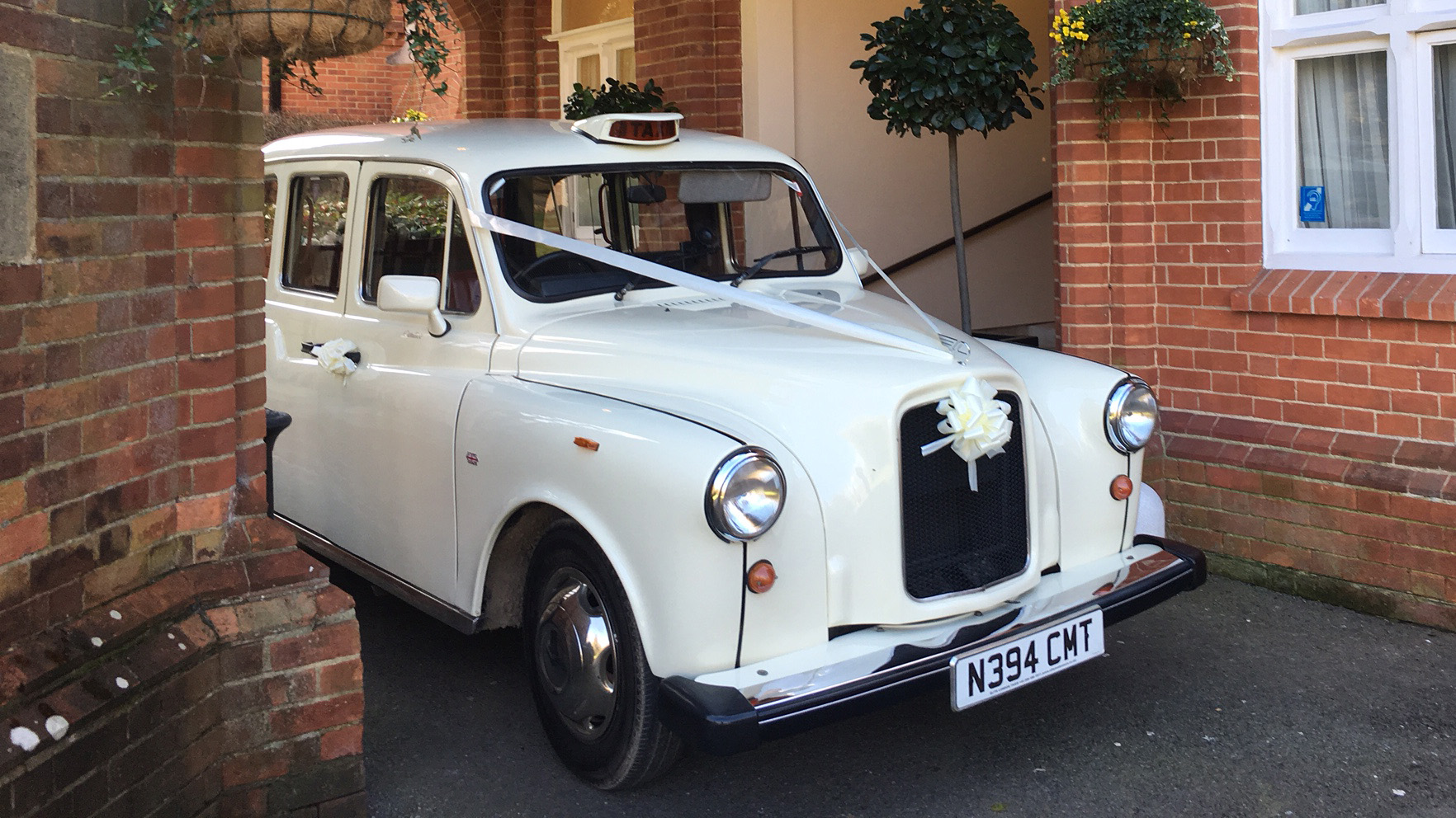 Old English White taxi cab decorated with ribbon and bow on the front, parked in a courtyard setting.