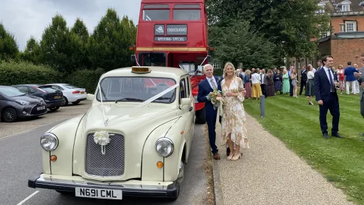 Classic ivory taxi cab arriving at a wedding with guests standing nearby, vehicle decorated with ribbons and parked on a gravel drive.