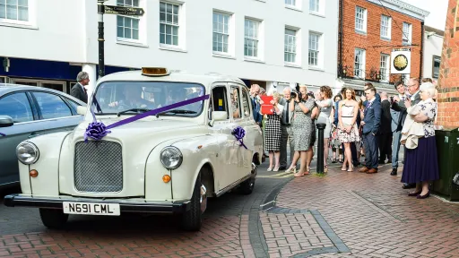 Old English White vintage taxi cab with purple wedding ribbons, parked outside a venue with guests gathered behind.