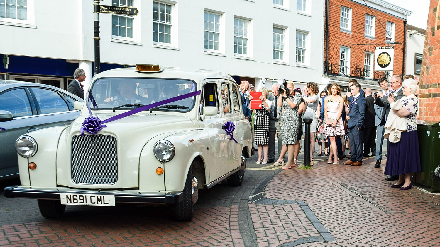 Old English White vintage taxi cab with purple wedding ribbons, parked outside a venue with guests gathered behind.