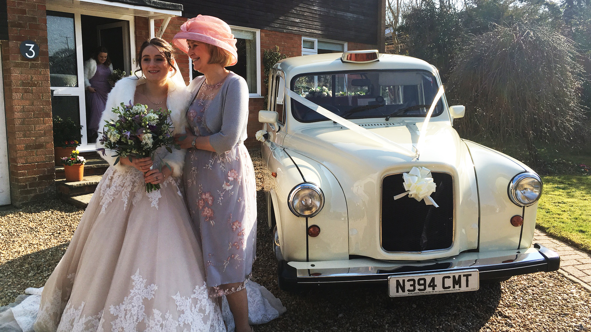 Mother and daughter standing beside a classic taxi cab at a wedding, vehicle decorated and parked outside a house.