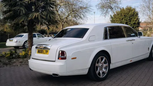 Rear three-quarter view of a white Rolls-Royce Phantom parked on a driveway with trees and residential background.