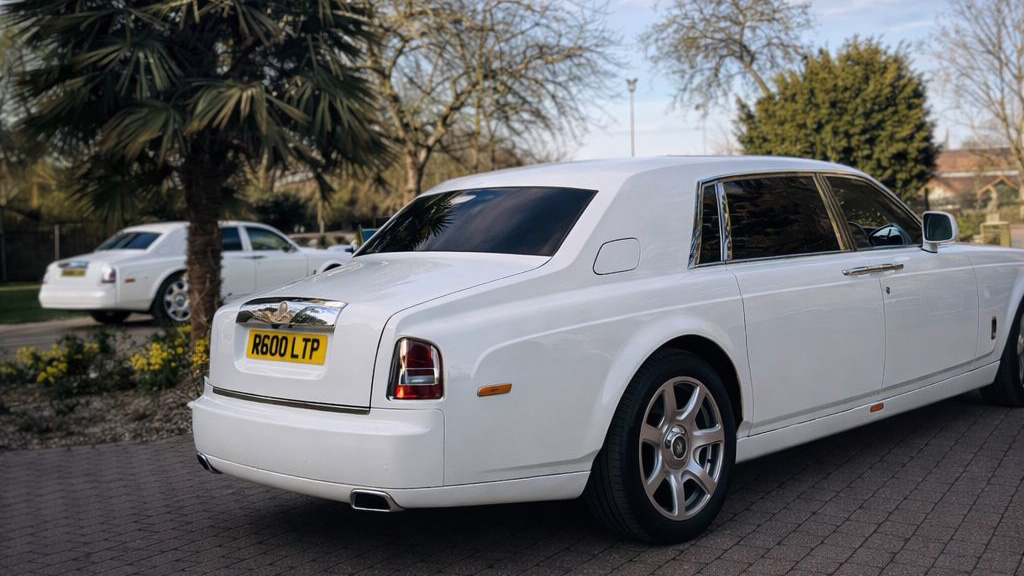 Rear three-quarter view of a white Rolls-Royce Phantom parked on a driveway with trees and residential background.