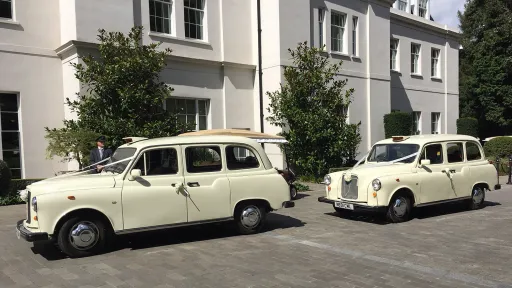 Two matching Old English White classic taxi cabs parked side by side outside a modern wedding venue, front angles visible with chrome grilles and clean driveway setting.