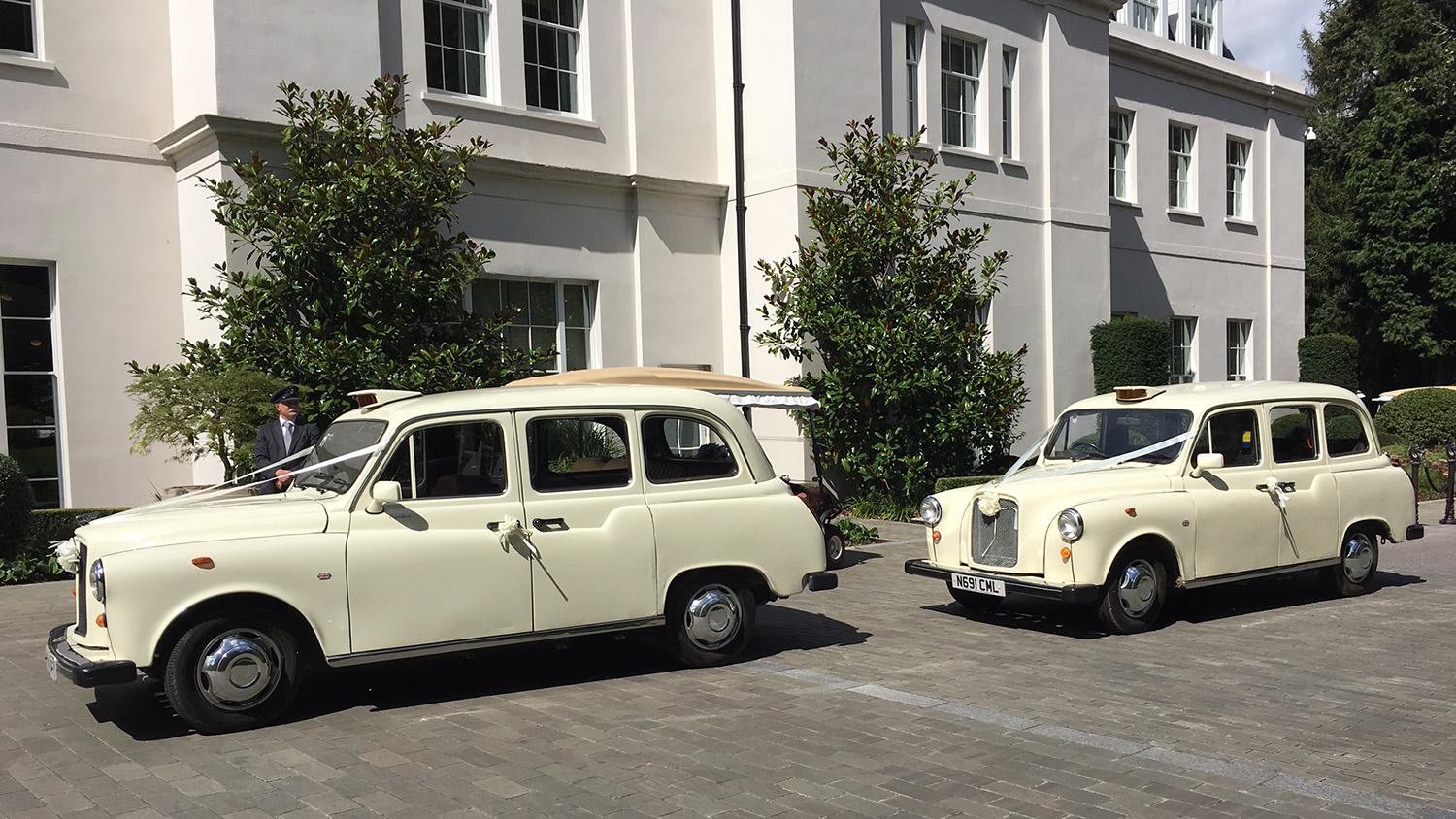 Two matching Old English White classic taxi cabs parked side by side outside a modern wedding venue, front angles visible with chrome grilles and clean driveway setting.