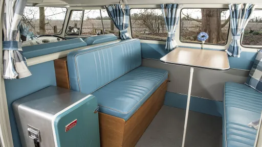 Rear seating area inside a VW campervan with turquoise upholstered seats, wooden cabinetry and spacious cabin layout.