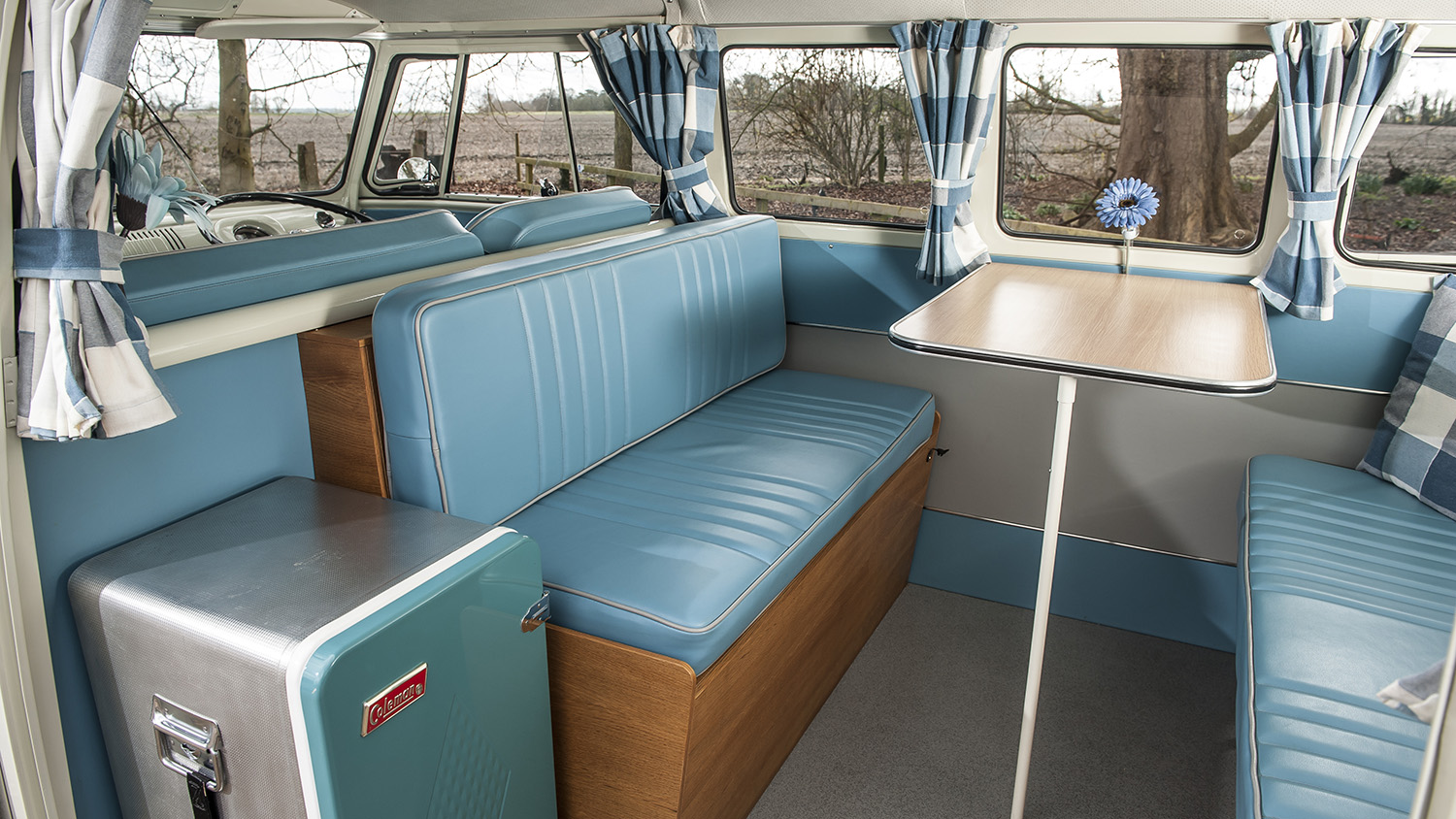 Rear seating area inside a VW campervan with turquoise upholstered seats, wooden cabinetry and spacious cabin layout.
