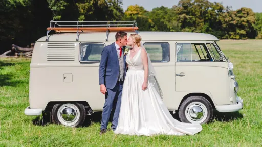 Bride and groom kissing beside a cream VW split screen campervan in a field, relaxed outdoor wedding setting.