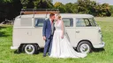 Bride and groom kissing beside a cream VW split screen campervan in a field, relaxed outdoor wedding setting.