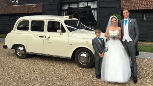 Bride and groom standing together with an Old English White taxi cab in the background, outdoor wedding setting.