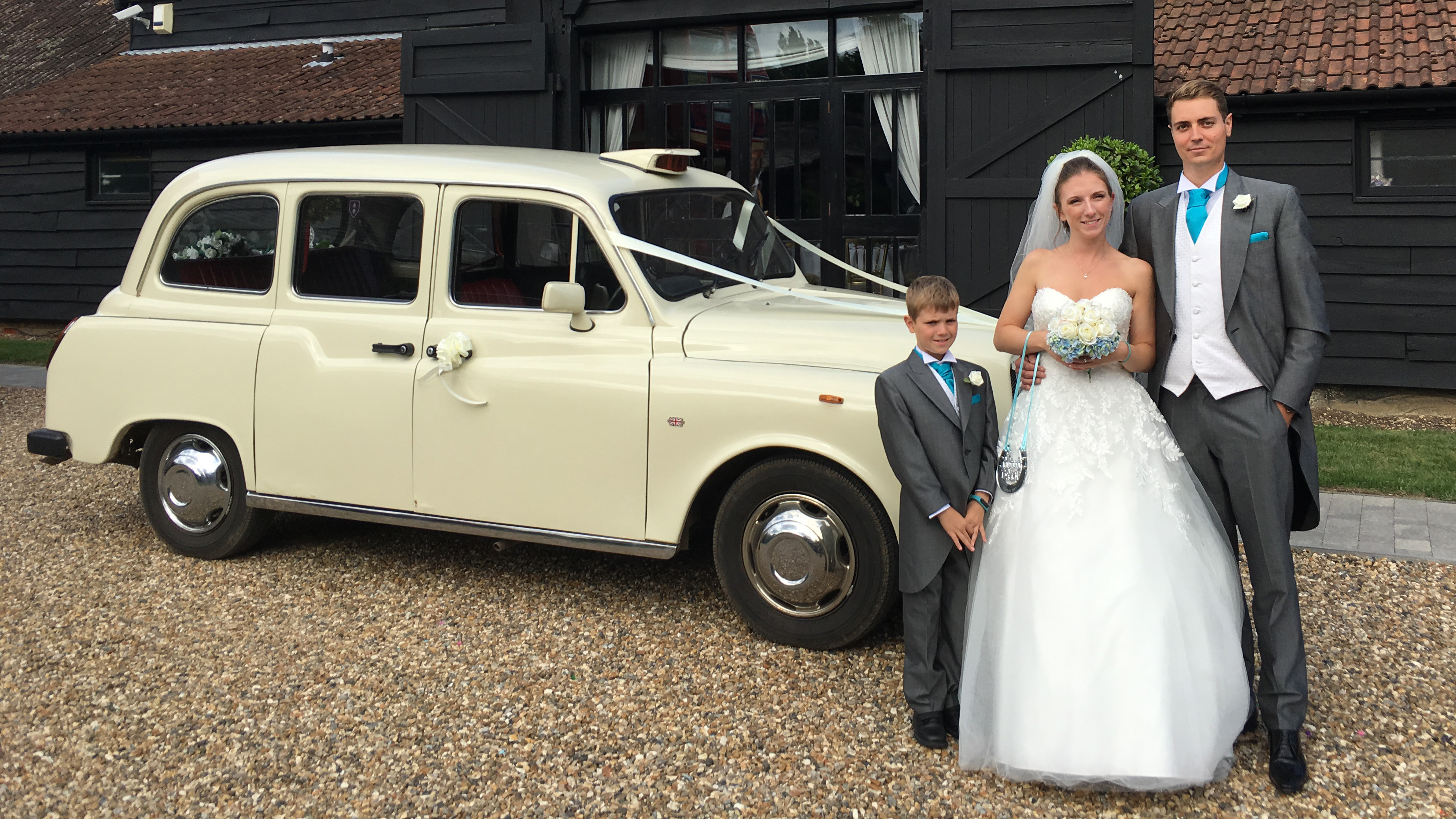 Bride and groom standing together with an Old English White taxi cab in the background, outdoor wedding setting.