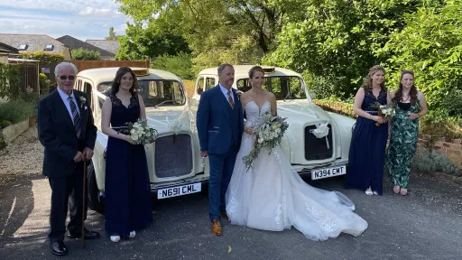 Bride and wedding party standing beside two identical ivory classic taxi cabs decorated with ribbons, parked outdoors with greenery and buildings in the background.