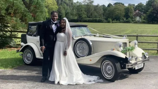 Bride and groom standing beside cream Beauford convertible wedding car with black soft top folded down, parked in countryside setting.