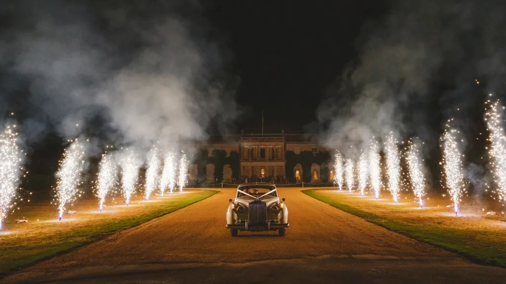 Classic Rolls-Royce Silver cloud exiting wedding venue at night with flairs on both side of the car