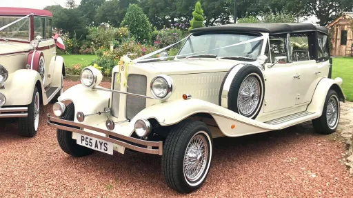 Cream Beauford 4 door convertible wedding car parked on a driveway, black soft top roof folded down, chrome detailing visible, countryside garden background.