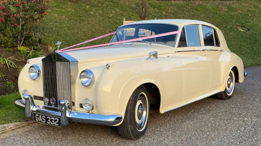 Front left view of Rolls-Royce Silver Cloud in Old English White with wedding ribbons on bonnet, parked near stone wall and greenery.