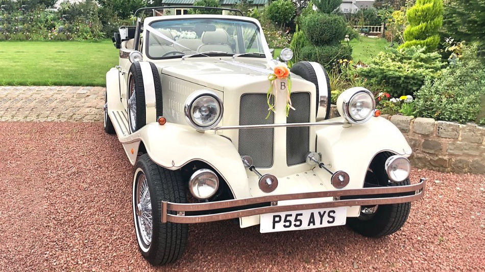 Front view of cream Beauford convertible wedding car with black folded roof, decorated with wedding ribbons on grille, parked on gravel drive with greenery behind.