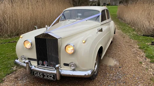 Front view of Rolls-Royce Silver Cloud in Old English White with iconic chrome grille and headlights, parked outdoors on gravel.