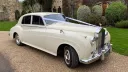 Side angle of Rolls-Royce Silver Cloud in Old English White parked on driveway, showing long body shape and classic styling.