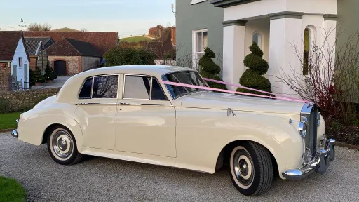 Rolls-Royce Silver Cloud in Old English White parked outside wedding venue with ribbons, buildings and trees in background.