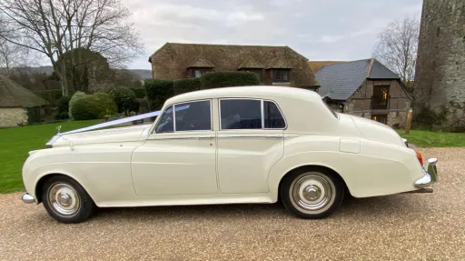 Full left side profile of Rolls-Royce Silver Cloud in Old English White parked on gravel with countryside and buildings behind.