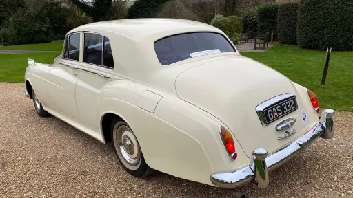 Rear left view of Rolls-Royce Silver Cloud in Old English White parked on gravel, showing rounded rear styling and chrome bumper.
