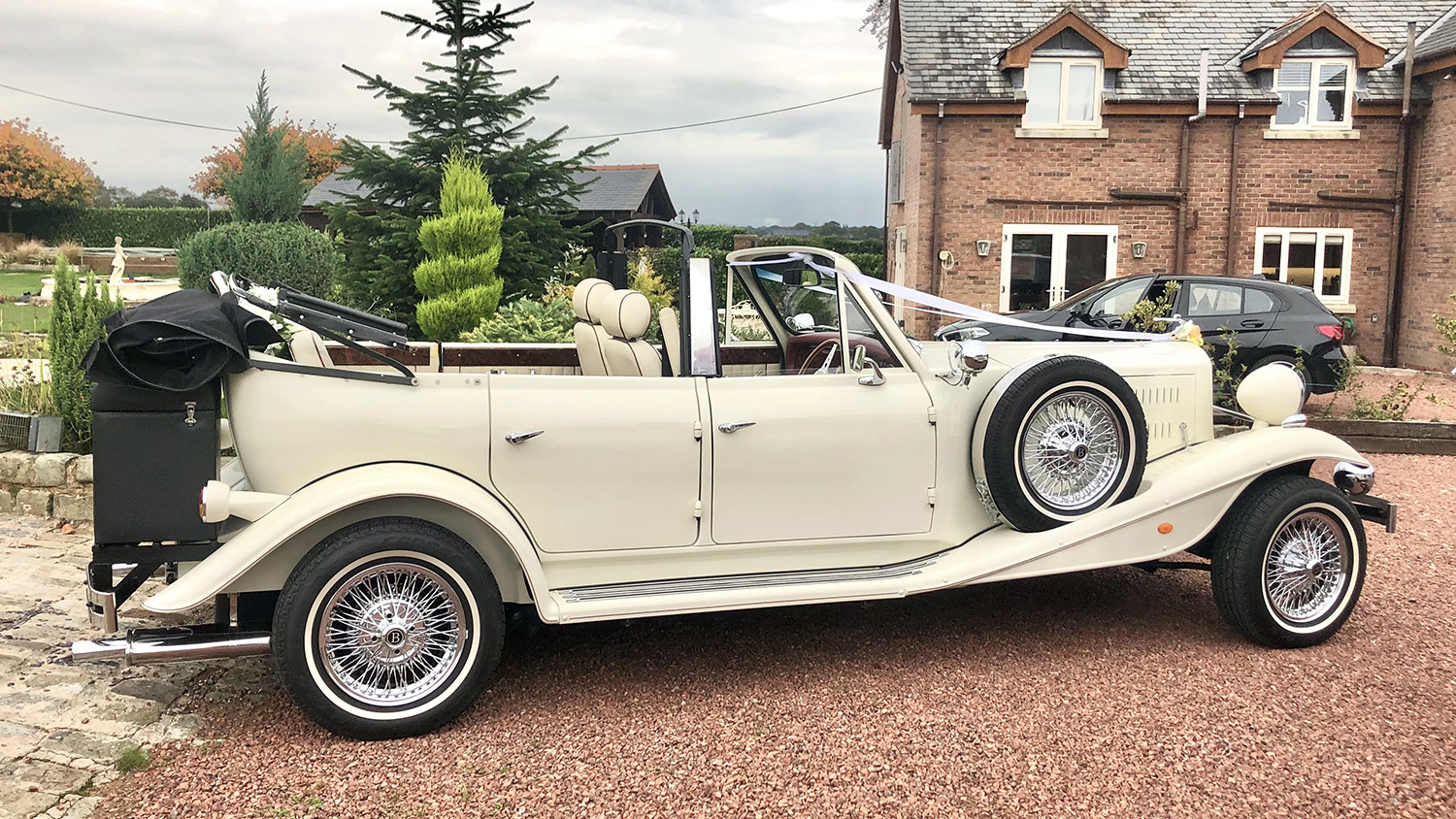 Side view of cream Beauford 4 door convertible with black soft top folded down, showing full length of vehicle, parked outside a house with brick driveway.