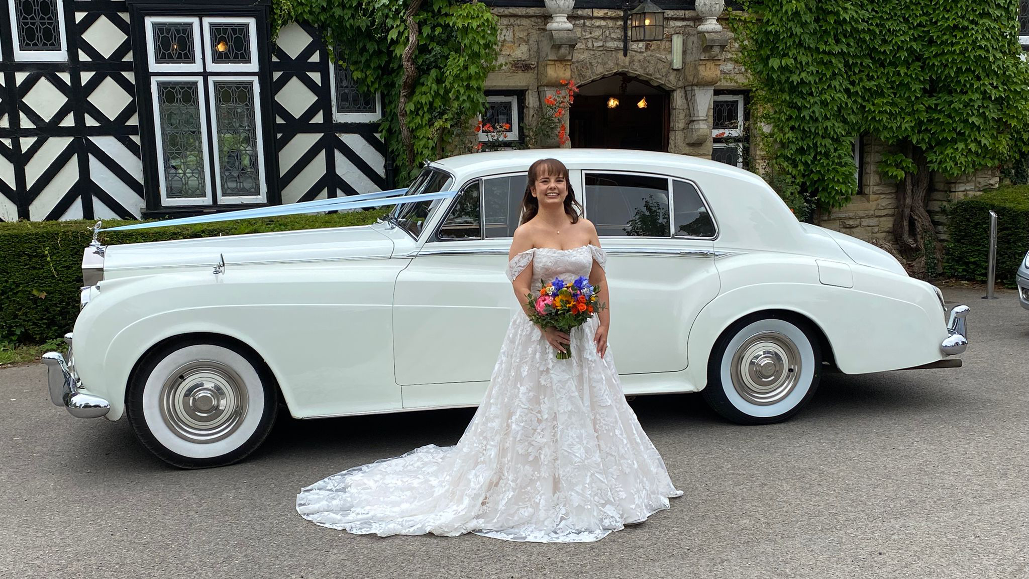 Bride in wedding dress standing beside 1957 Rolls-Royce Silver Cloud in Old English White outside traditional venue