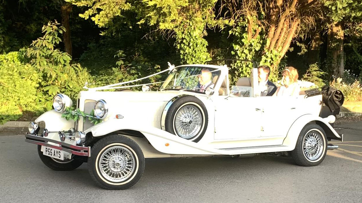 Cream Beauford 4 door convertible wedding car decorated with flowers and ribbons, parked on country road with black soft top folded down.