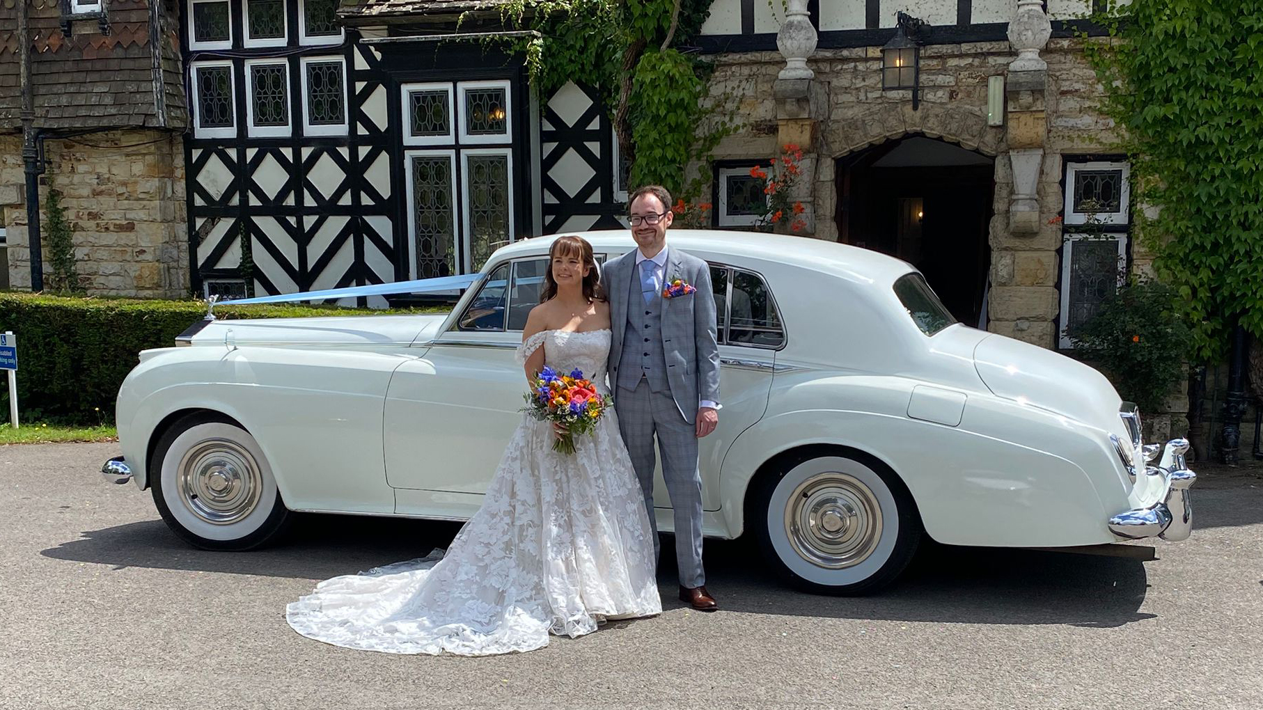 Bride and groom posing next to Rolls-Royce Silver Cloud in Old English White, historic building background