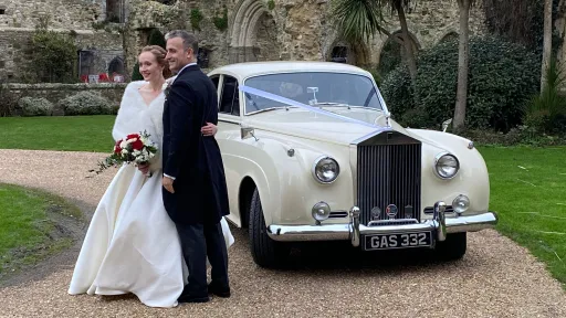 Bride and groom standing beside Rolls-Royce Silver Cloud in Old English White, parked on gravel in garden setting.