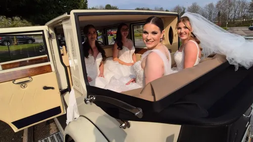 Bride and bridesmaids seated inside cream 1930s style Imperial car with roof open, showing cream leather interior and black soft top folded back.