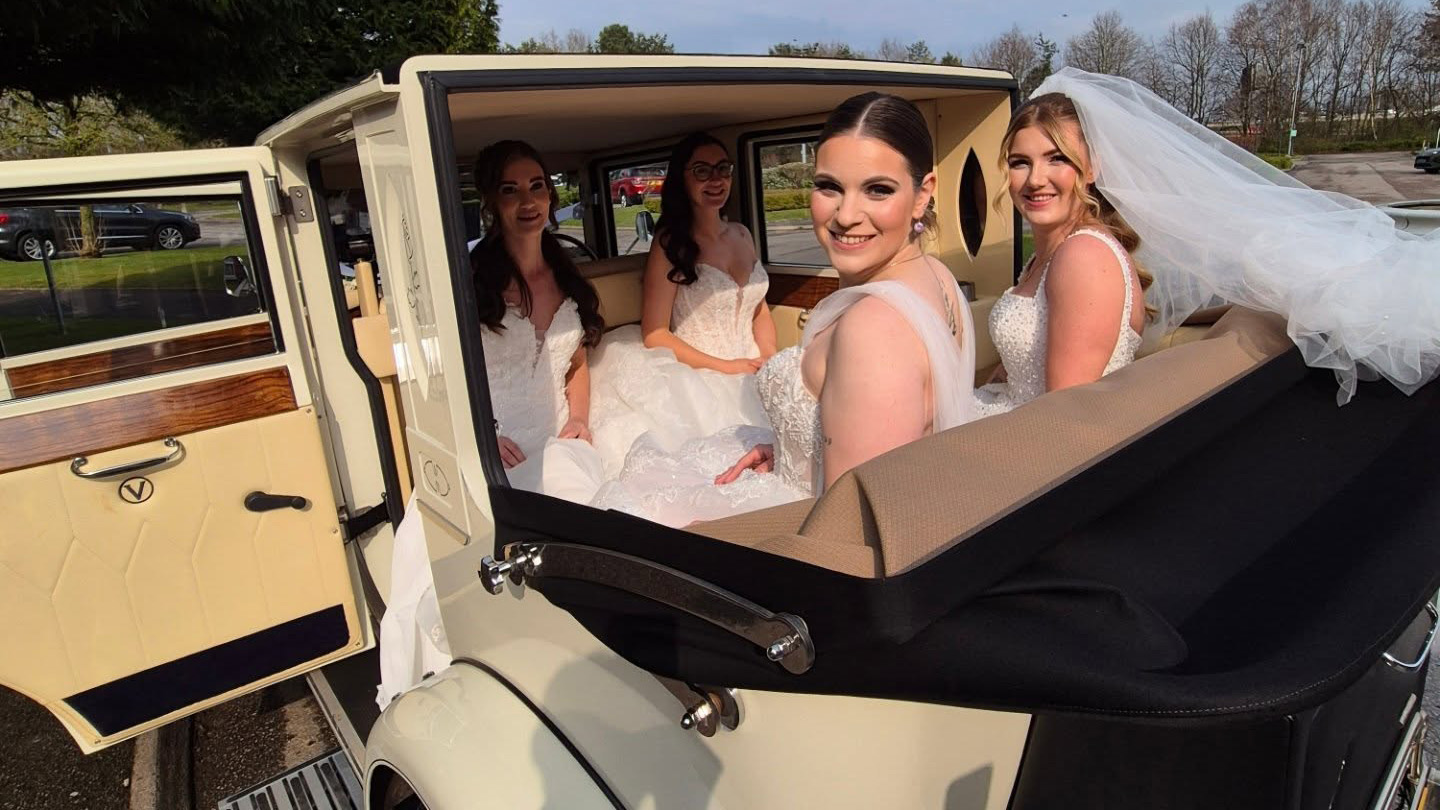 Bride and bridesmaids seated inside cream 1930s style Imperial car with roof open, showing cream leather interior and black soft top folded back.