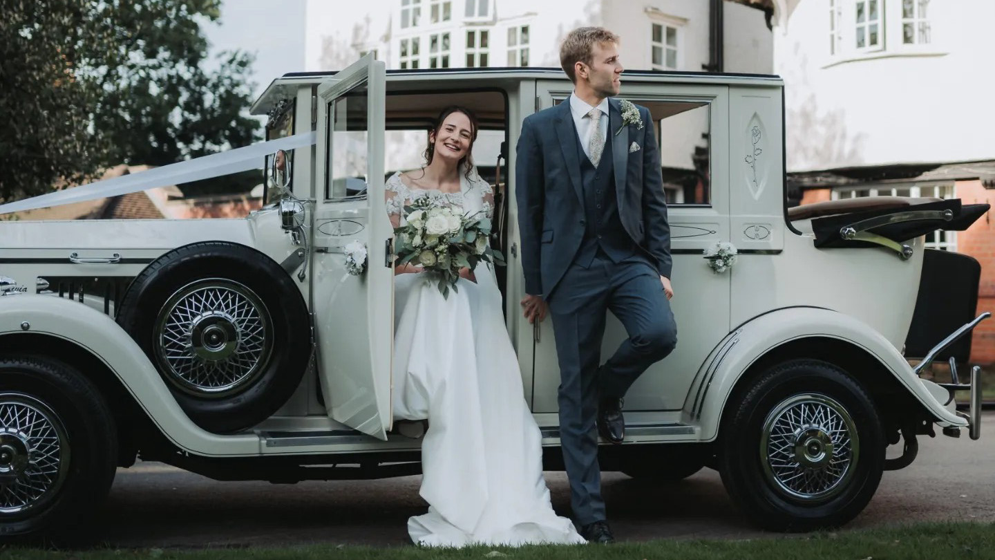 Bride and groom posing beside a cream vintage Imperial wedding car with chrome details and black soft top roof, parked outside a venue.