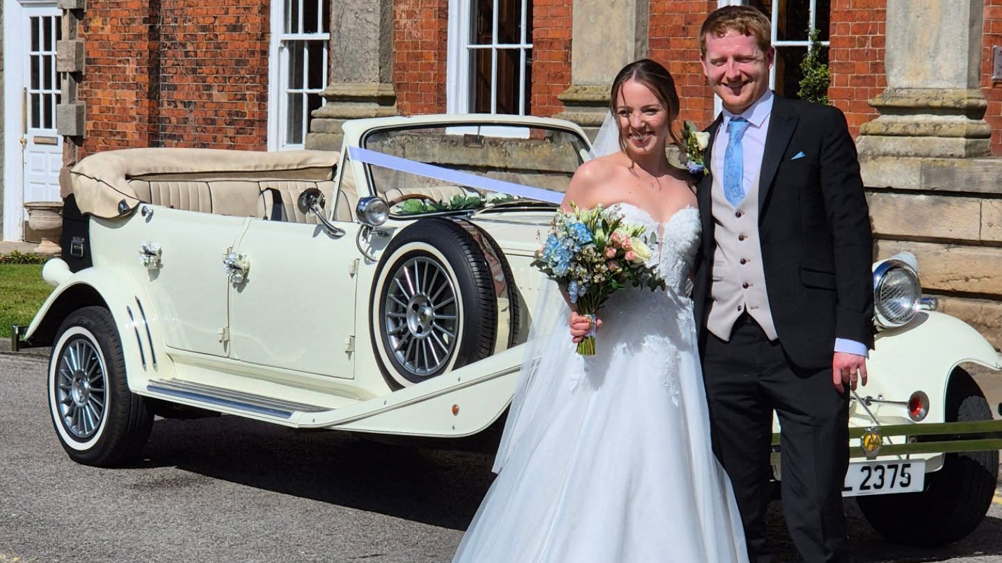 Bride and groom posing next to cream Beauford convertible wedding car at a venue in Warrington, Cheshire.