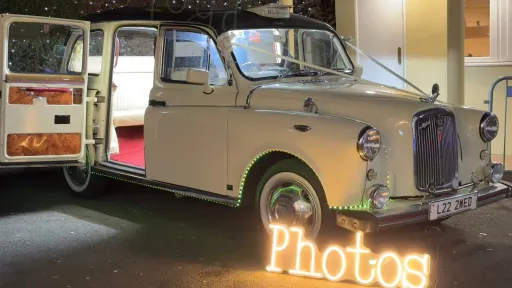 Classic ivory taxi cab converted with built-in photobooth, rear doors open showing burgundy carpet and interior setup.