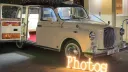 Classic ivory taxi cab converted with built-in photobooth, rear doors open showing burgundy carpet and interior setup.