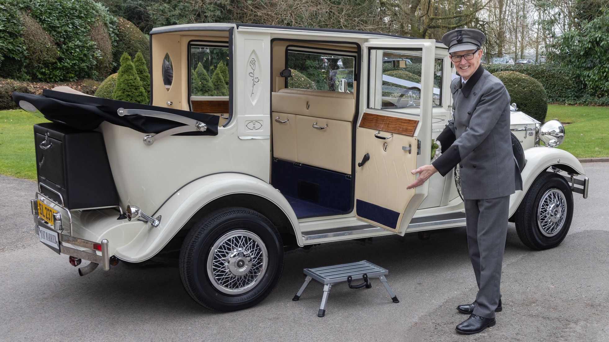 Uniformed chauffeur opening rear door of cream Imperial wedding car with black soft top roof and chrome detailing.