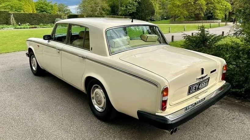 Rear left view of a cream Rolls-Royce Silver Shadow Mk2 parked on a quiet road with greenery in the background.