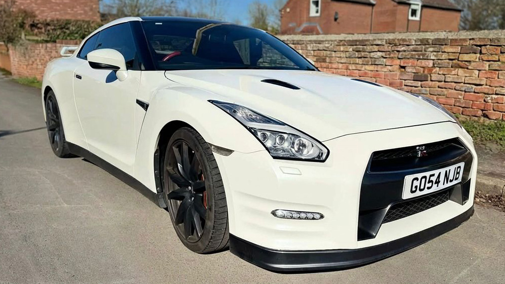 White Nissan GTR R35 wedding car parked at angle, sleek coupe design with black alloy wheels, set against brick wall and driveway.