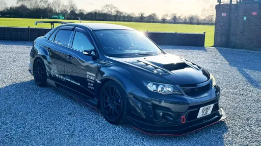 Black Subaru Impreza performance car parked on gravel, front three-quarter angle, aggressive styling with bonnet scoop and sporty body kit, countryside background.