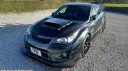Black Subaru Impreza wedding hire car viewed from front angle, bonnet scoop and low stance visible, parked on open gravel area with rural surroundings.