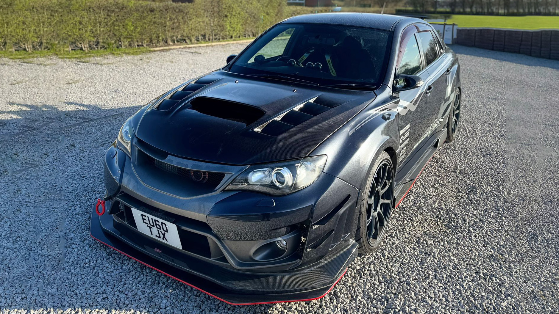 Black Subaru Impreza wedding hire car viewed from front angle, bonnet scoop and low stance visible, parked on open gravel area with rural surroundings.