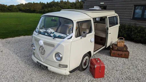 Classic Volkswagen Campervan bay window dressed with white ribbons and floral decoration, and period suitcases outside open rear door
