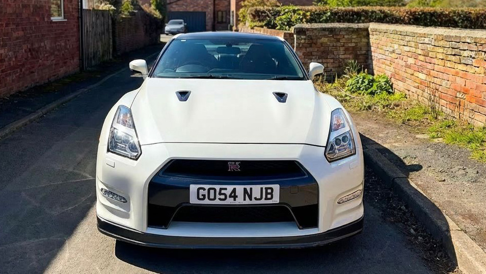 Front view of white Nissan GTR R35 parked on driveway, aggressive grille and bonnet lines visible, residential brick wall background.
